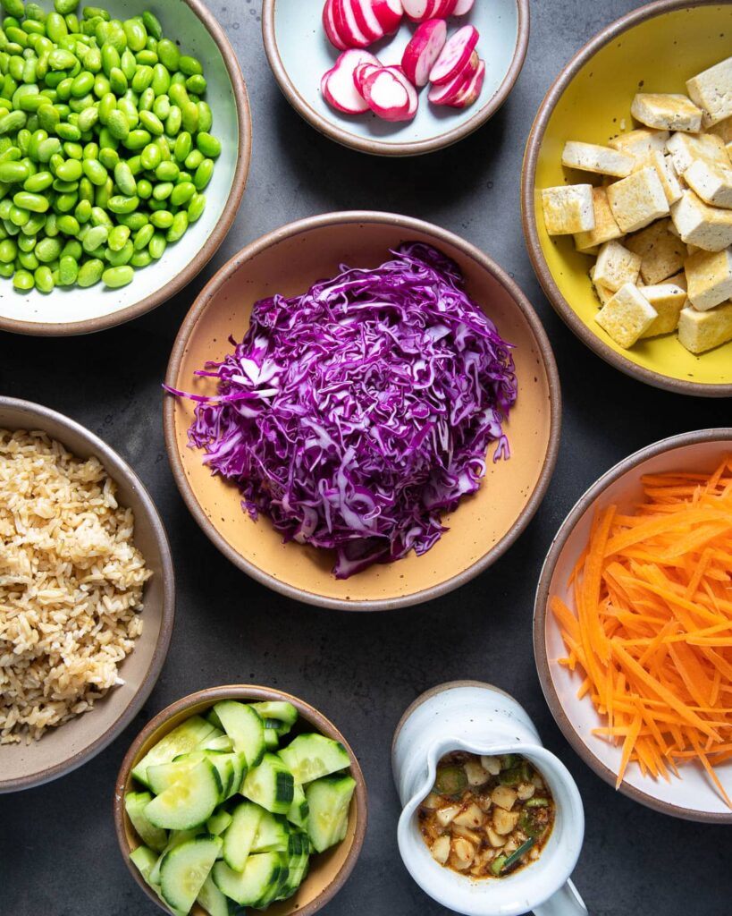 poke bowl ingredients, edamame, radishes, red cabbage. tofu, carrots, rice, cucumbers, sauce